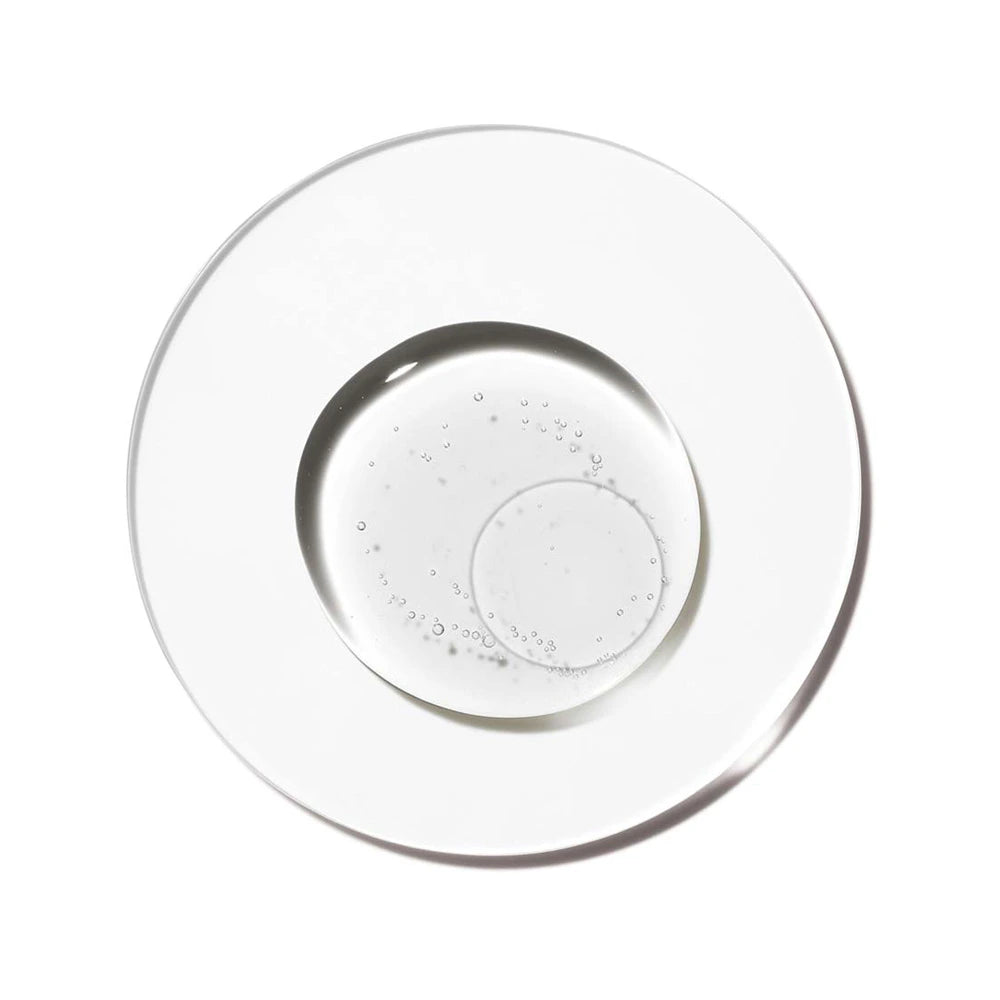 A top-down, minimalist laboratory shot showcasing pure hemisqualane fluid inside a clear glass Petri dish set against a stark white background. The transparent, lightweight emollient forms a perfectly circular puddle in the center, filled with numerous tiny, delicate air micro-bubbles suspended within the fluid. A secondary clear circular outline overlays the texture, accentuating the clean, scientific, and hydrating nature of the ingredient.