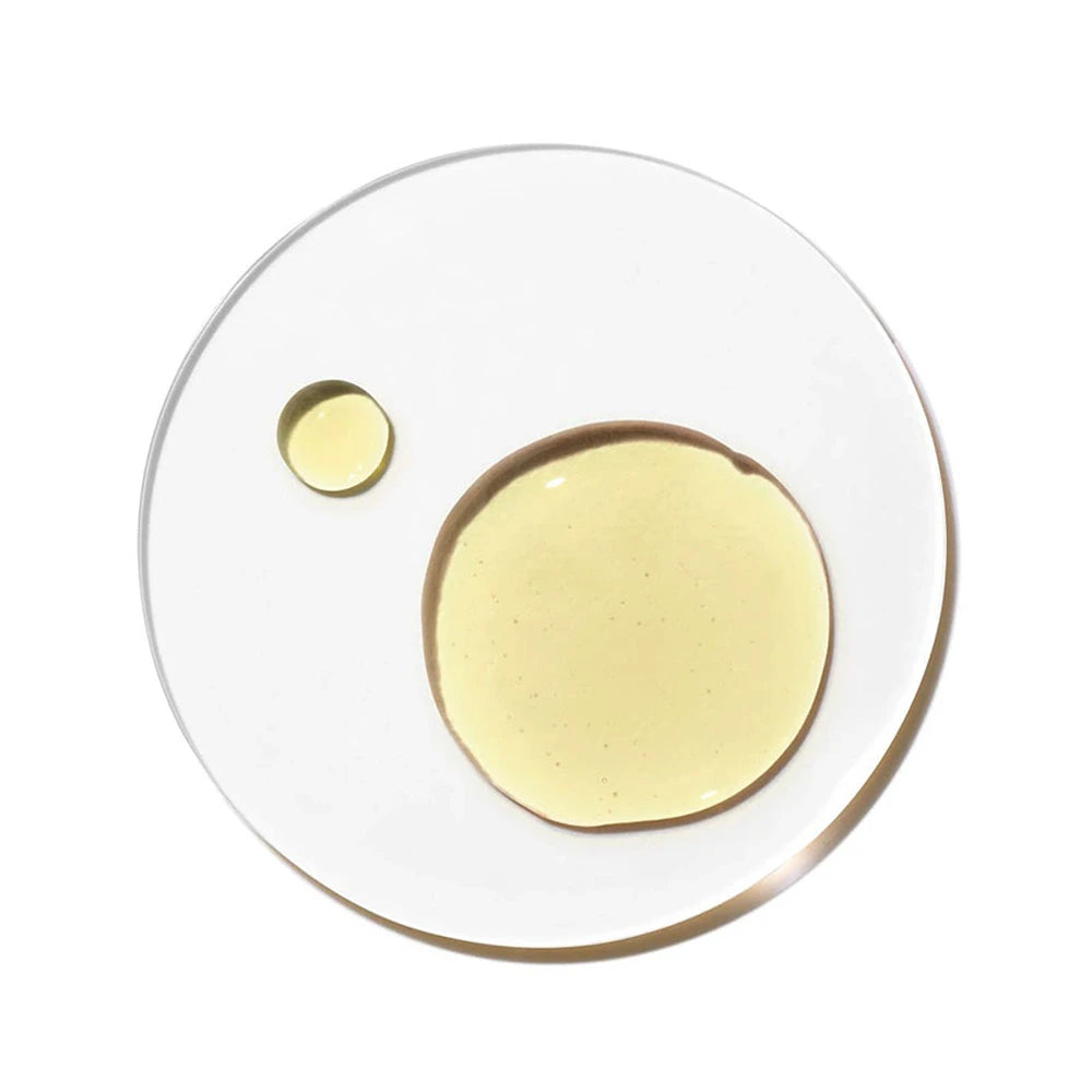 A top-down, minimalist laboratory-style cosmetic photograph featuring pure jojoba oil inside a perfectly circular, clear glass Petri dish against a crisp white background. The image displays a smooth, rounded puddle of translucent golden-yellow oil alongside a smaller, separate circular droplet. The texture of the oil is clean and viscous, reflecting subtle light and cast shadows that emphasize its pure, nourishing quality.