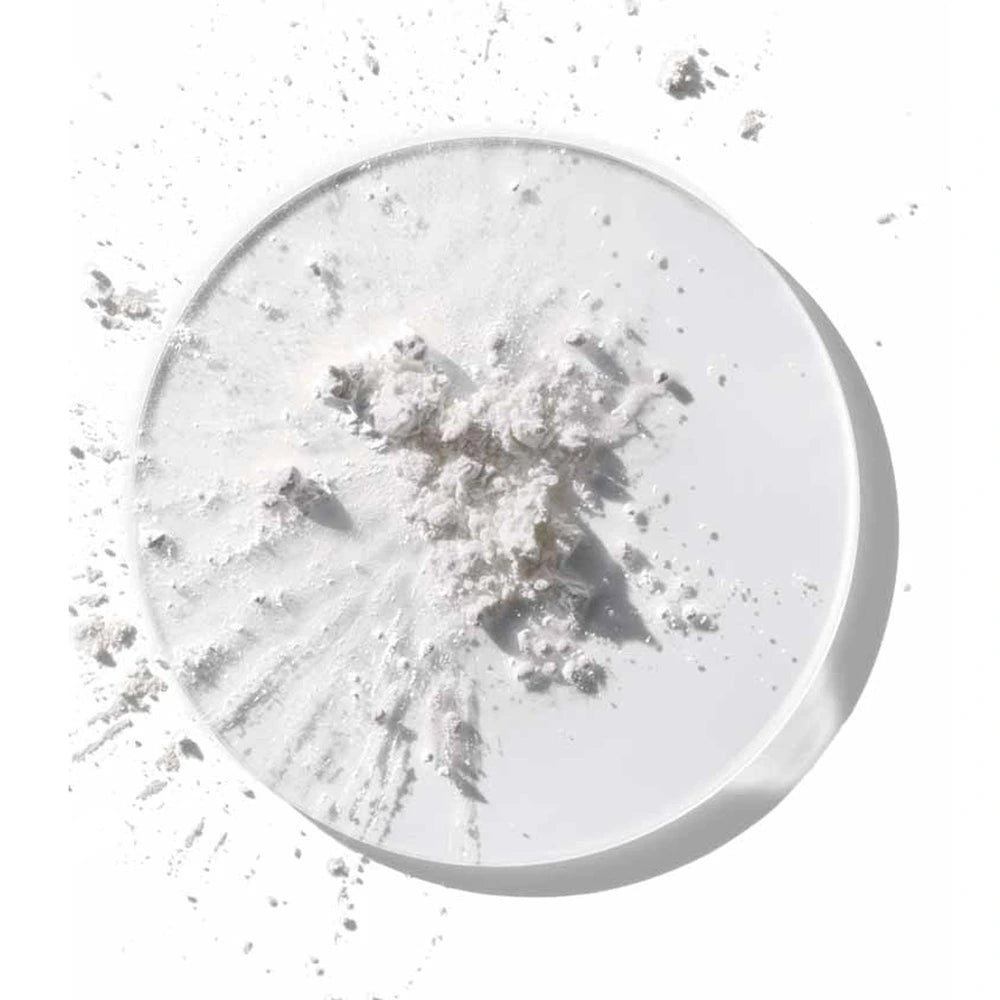 A high-angle, minimalist cosmetic ingredient shot showing fine white active powder scattered across and inside a round, clear glass Petri dish. The raw powder ingredient, representing peptides and ceramides, forms a small textured cluster in the center with delicate bursts and speckles radiating outward onto a clean, bright white background under soft, natural lighting.
