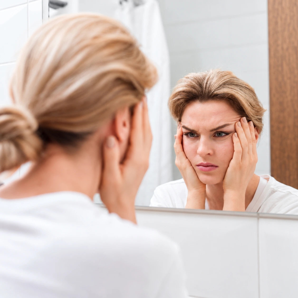 Blonde woman looking at her reflection in the mirror, checking for signs of aging on her skin.